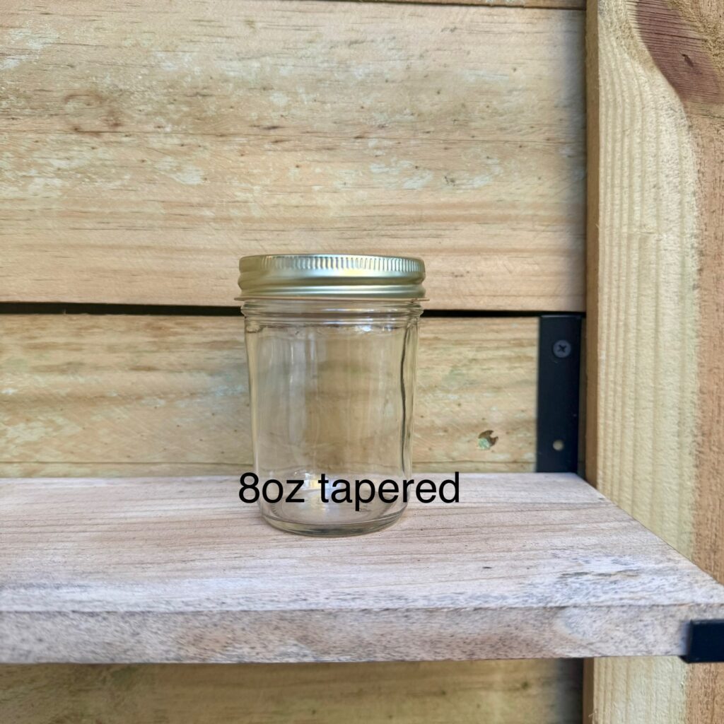 An 8oz tapered clear glass jar with metal lid on wooden shelf.